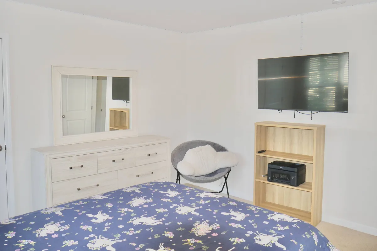 A bedroom with floral patterned bedding, a dresser with a mirror, a wall-mounted television, and a wooden shelf unit. The setting is cozy and welcoming, with soft lighting that conveys a restful and inviting mood.