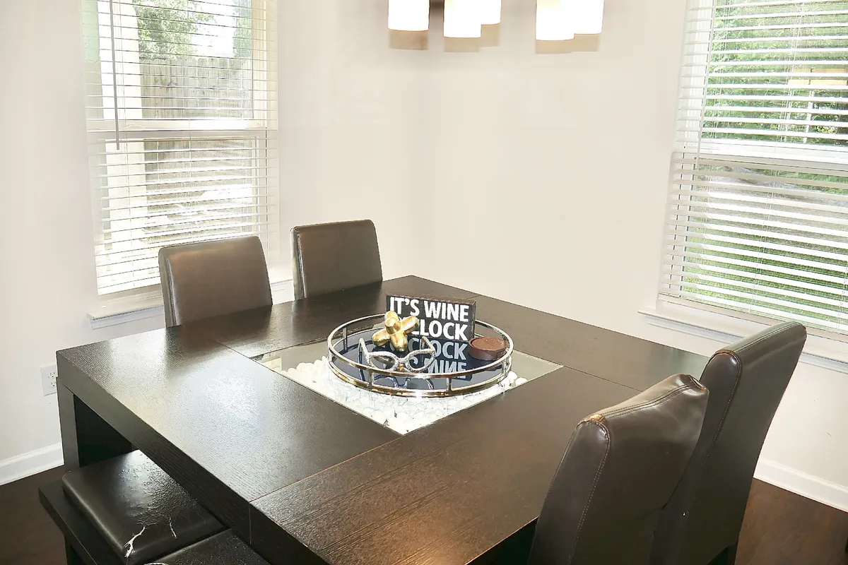 A warmly-lit dining area featuring a large table with a decorative centerpiece, surrounded by comfortable chairs and topped with a stylish light fixture. The setup suggests a space perfect for shared meals, celebrations, or game nights with friends and family.