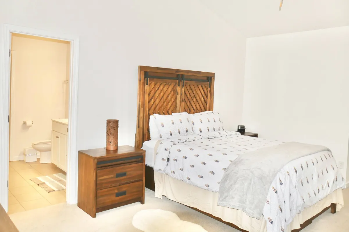 A serene bedroom with a decorative headboard, nightstand and lamp, and an open doorway that reveals a modern bathroom. The room has soft natural light and a sense of comfort, making it ideal for restful sleep or peaceful mornings.