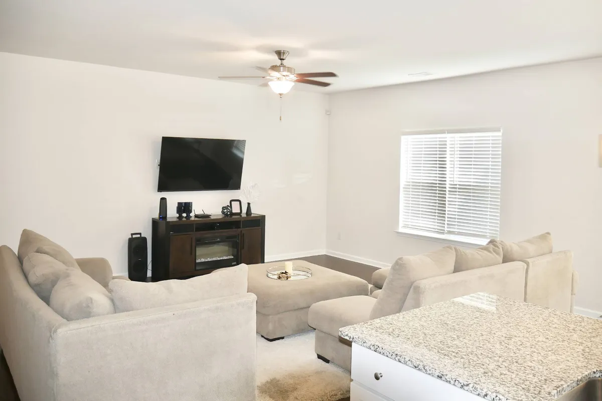 The hero image showcases a spacious, sunlit living room with a large, inviting sectional sofa arranged around a sleek coffee table, facing a wall-mounted TV. Ample windows with blinds bathe the space in natural light, creating a warm and welcoming atmosphere perfect for gathering with friends or family.