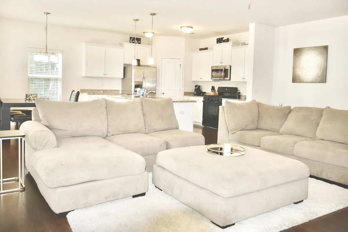 A spacious, open-concept living area featuring a large sectional sofa arranged around a coffee table, with the kitchen island visible in the background. The room exudes a cozy, welcoming ambiance, ideal for relaxation, socializing, and family gatherings.
