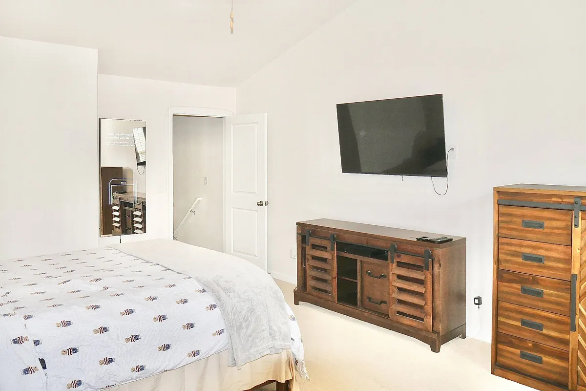 A bedroom with a comfortable bed featuring patterned bedding, a wooden dresser, and a wall-mounted television. Natural light fills the space, creating a relaxing environment ideal for unwinding after a day of work or travel.