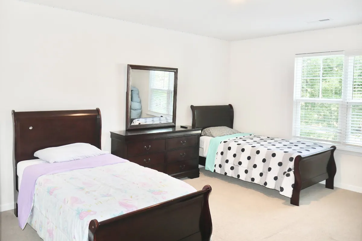 A welcoming bedroom showcasing two comfortable beds, a dresser, mirror, and a window that bathes the room in morning sunlight. The space feels cozy, ideal for families, teens, or friends traveling together.